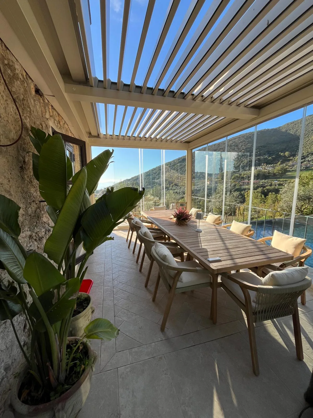 Tavolo apparecchiato sotto una pergola ECCELSA, lamelle chiuse e piante ai lati