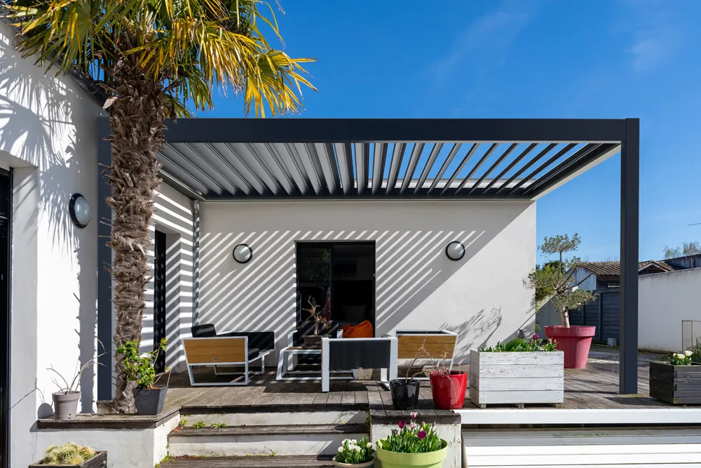 Pergola ESSENZA antracite addossata a una casa bianca, le lamelle orientabili aperte